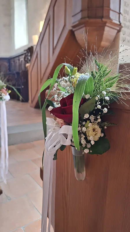 Rote Rose, helle Blüten und weisse Bänder als Dekoration an einer Kirchenbank